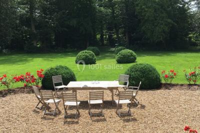 Table et chaises en bois disposées sur du gravier, face à un jardin verdoyant avec buissons et pelouse.