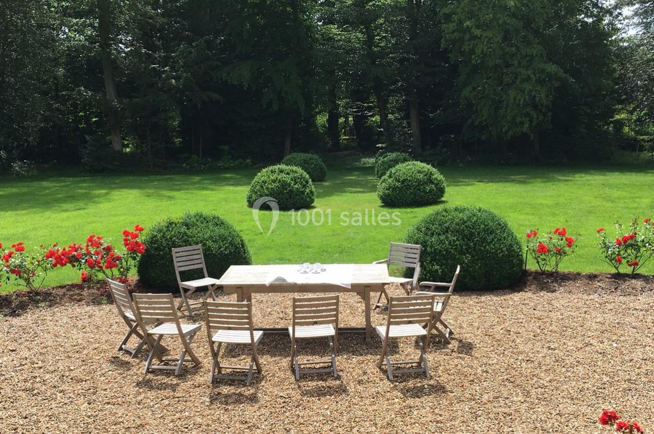 Table et chaises en bois disposées sur du gravier, face à un jardin verdoyant avec buissons et pelouse.