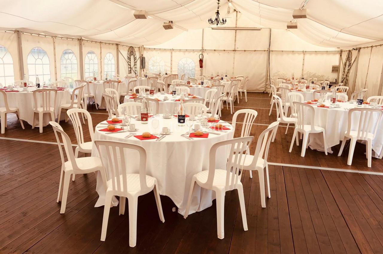 Salle de réception avec tables rondes dressées, nappes blanches, chaises en plastique et décoration sobre sous une tente.