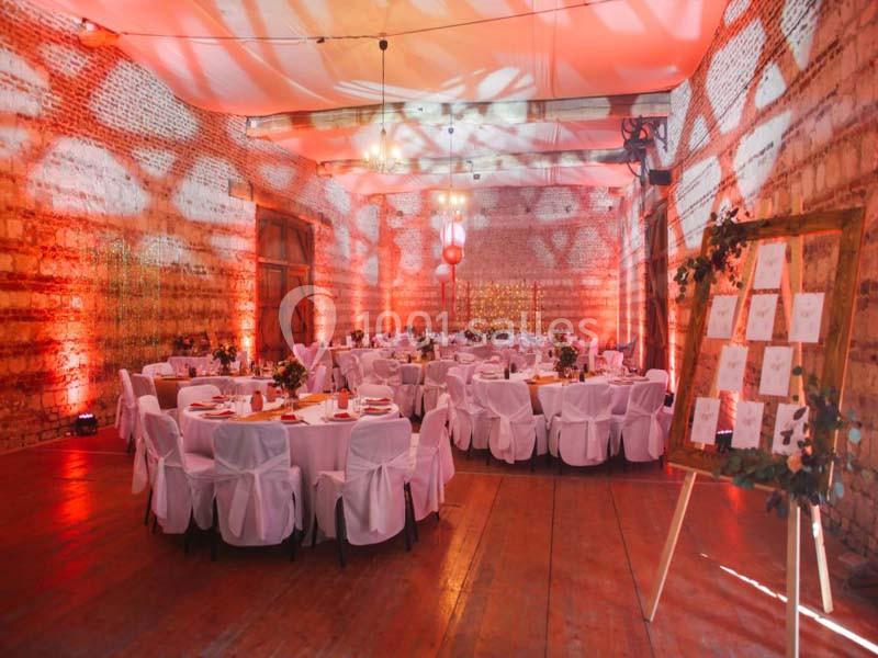Salle de réception décorée avec des tables rondes, nappes blanches et éclairage chaleureux aux tons rouges.