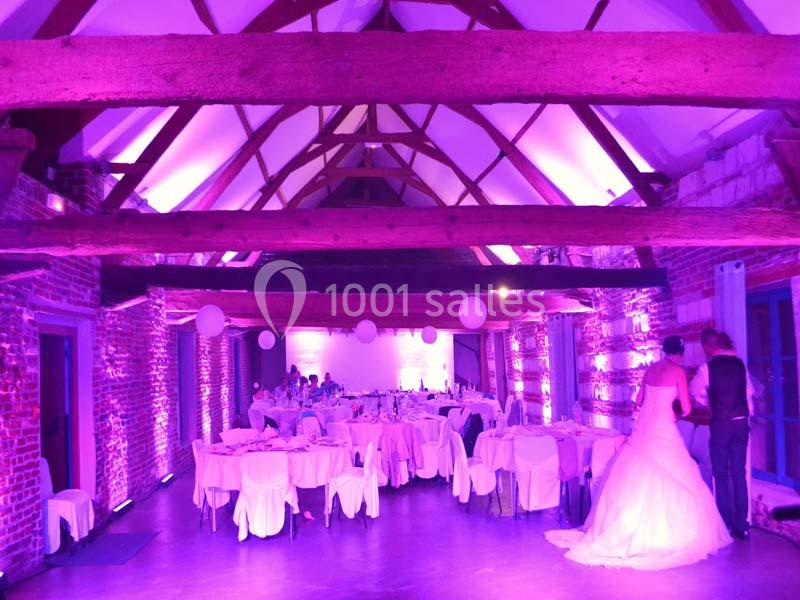 Salle de réception avec poutres apparentes, tables dressées et éclairage violet, un couple en tenue de mariage au fond.
