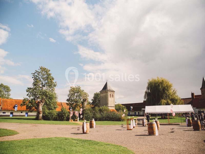 Cour extérieure avec pelouse, tonneaux en bois, tente blanche et bâtiments en briques sous un ciel partiellement nuageux.