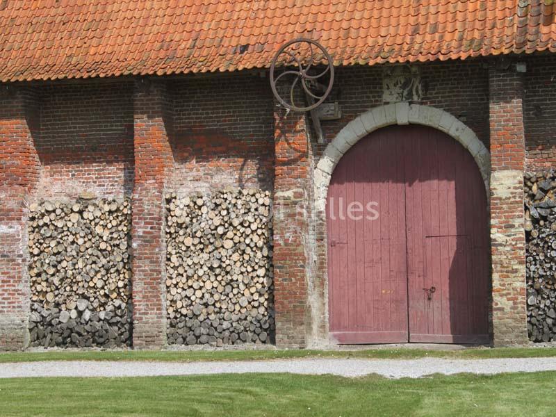 Façade en briques d'une grange avec porte en bois rouge, toit en tuiles et piles de bois empilées sur les côtés.