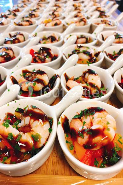 Petites coupelles blanches contenant des crevettes, légumes et sauce balsamique, disposées en rangées sur une table.