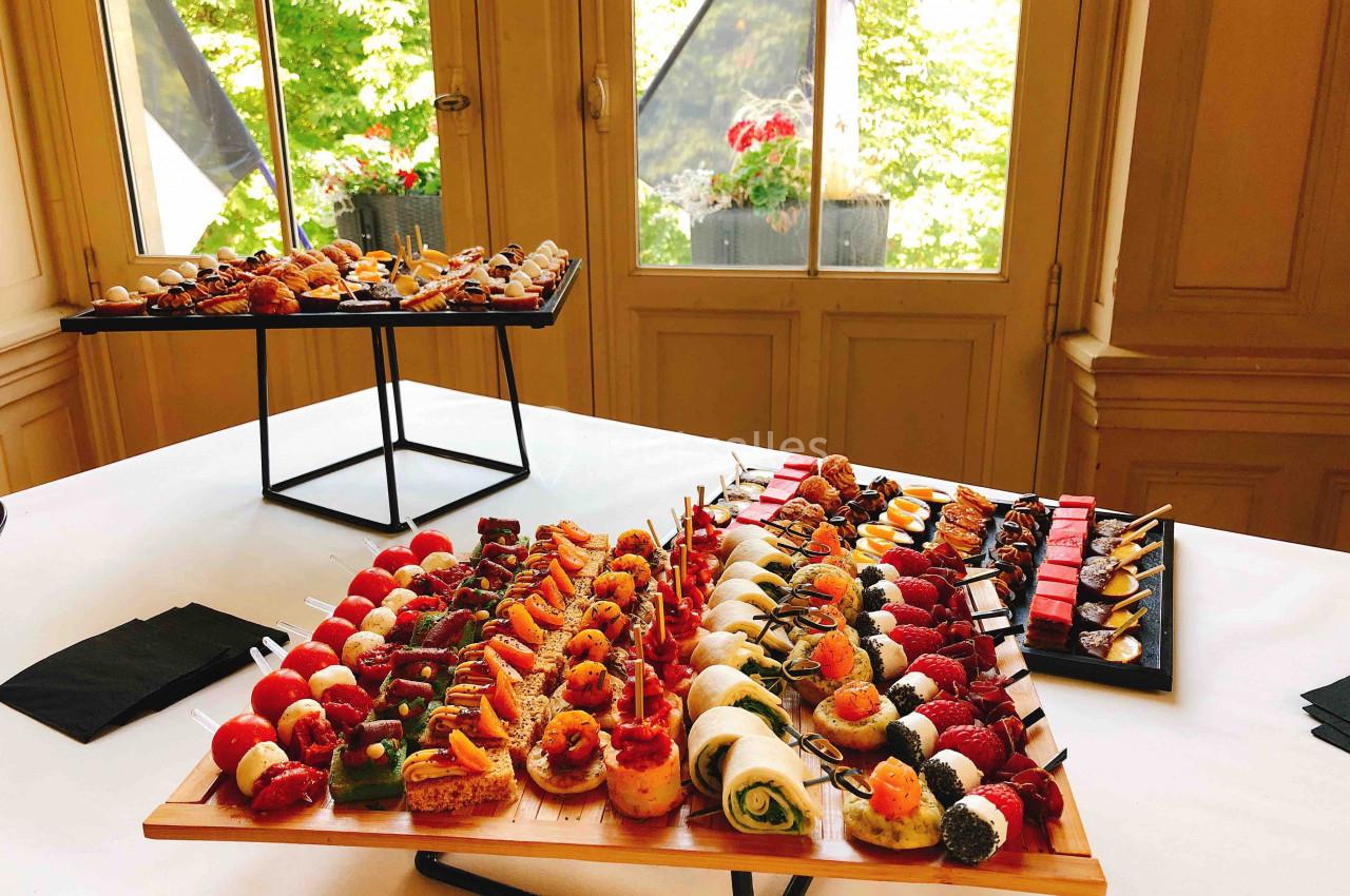 Plateaux de bouchées apéritives variées disposés sur une table blanche dans une salle lumineuse avec vue sur un jardin.