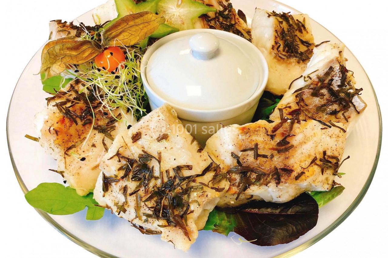 Assiette de poisson grillé aux herbes, accompagnée de légumes frais, d'une sauce en ramequin et de garnitures décoratives.