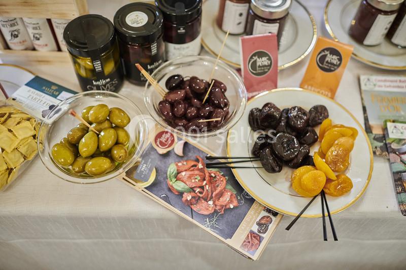 Assortiment d'olives, fruits secs et confitures disposés sur une table avec brochures en arrière-plan.