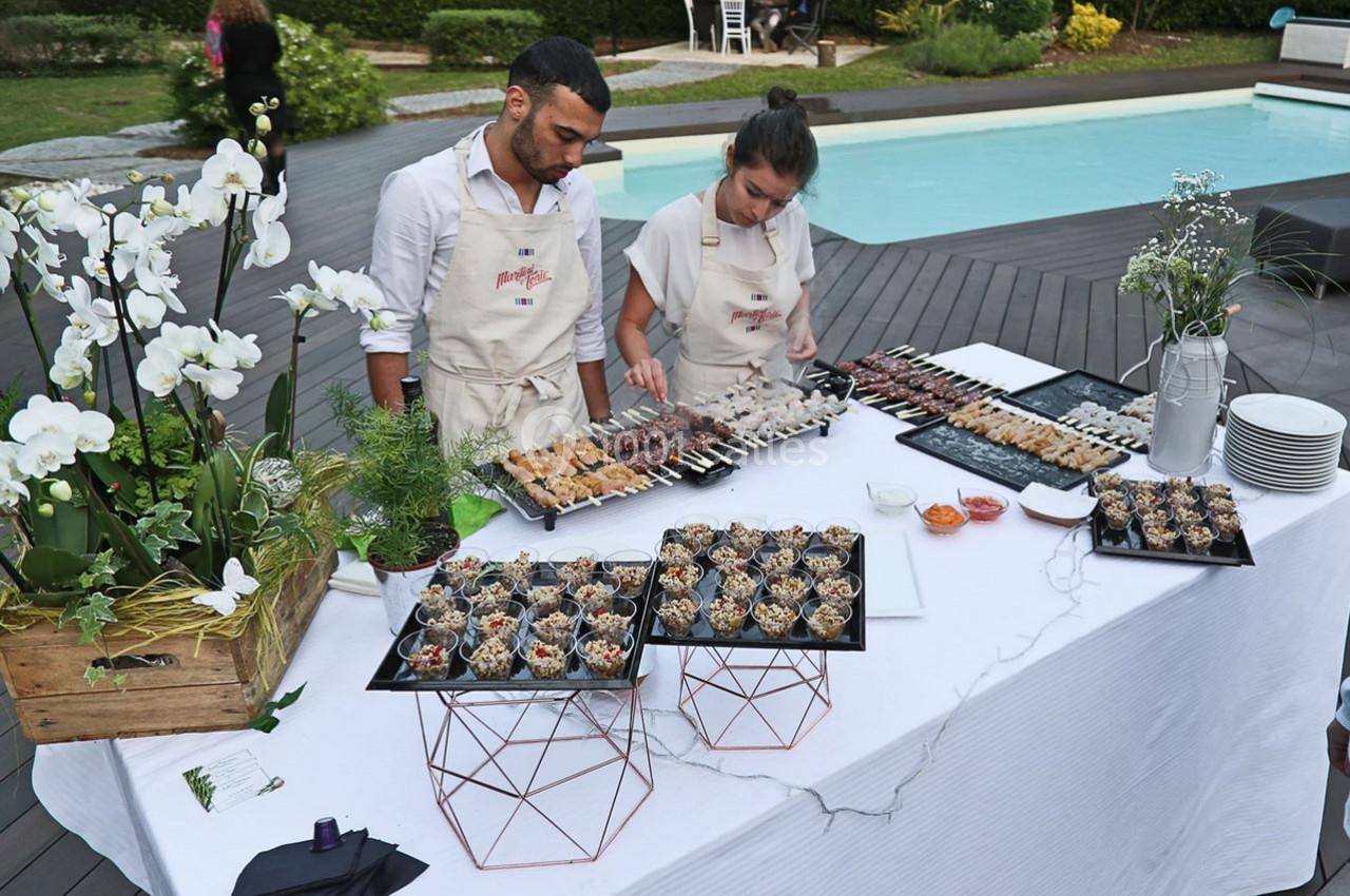 Deux personnes préparent un buffet de brochettes et verrines sur une table dressée près d'une piscine.