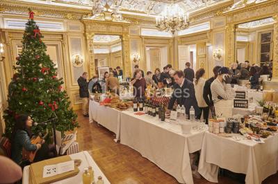Une femme souriante se tient derrière un stand de produits variés dans une salle décorée avec un sapin de Noël.