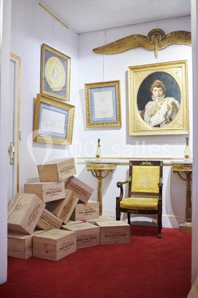 Des caisses en bois empilées dans une pièce ornée de cadres dorés, d'un fauteuil et d'un portrait impérial.