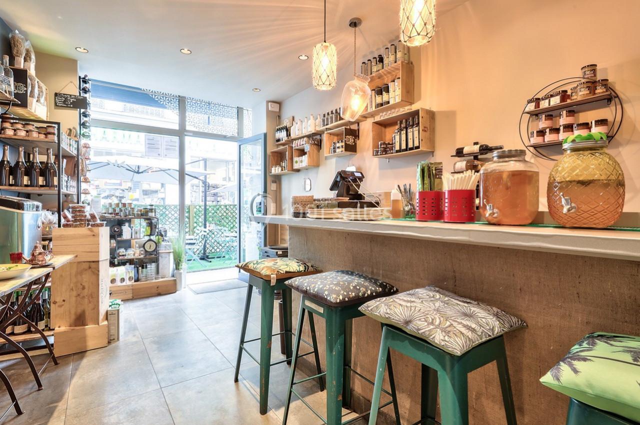 Intérieur d'un café chaleureux avec comptoir, tabourets colorés, étagères garnies et vue sur une terrasse extérieure.