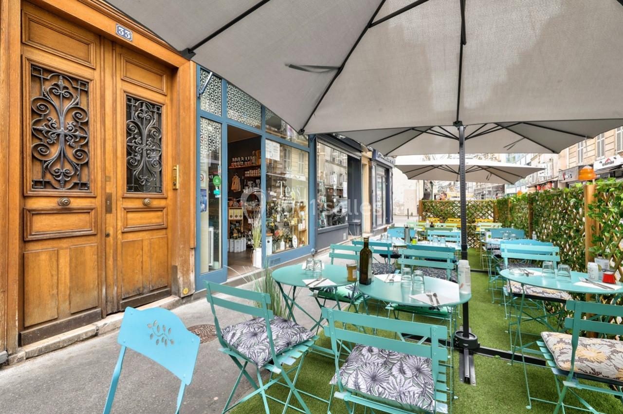Terrasse extérieure avec tables et chaises turquoise, parasols, et vue sur une devanture de boutique en ville.