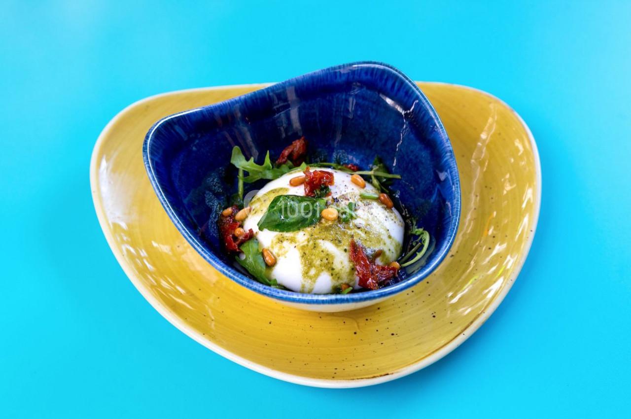 Assiette colorée contenant une burrata garnie de pesto, tomates séchées, pignons de pin et roquette.