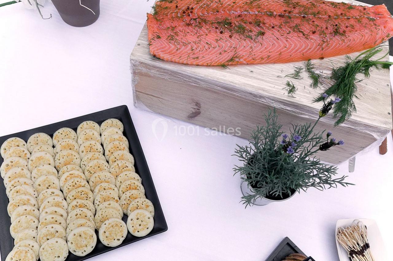 Saumon fumé sur planche en bois avec des blinis disposés sur une assiette noire, décoré de plantes aromatiques.