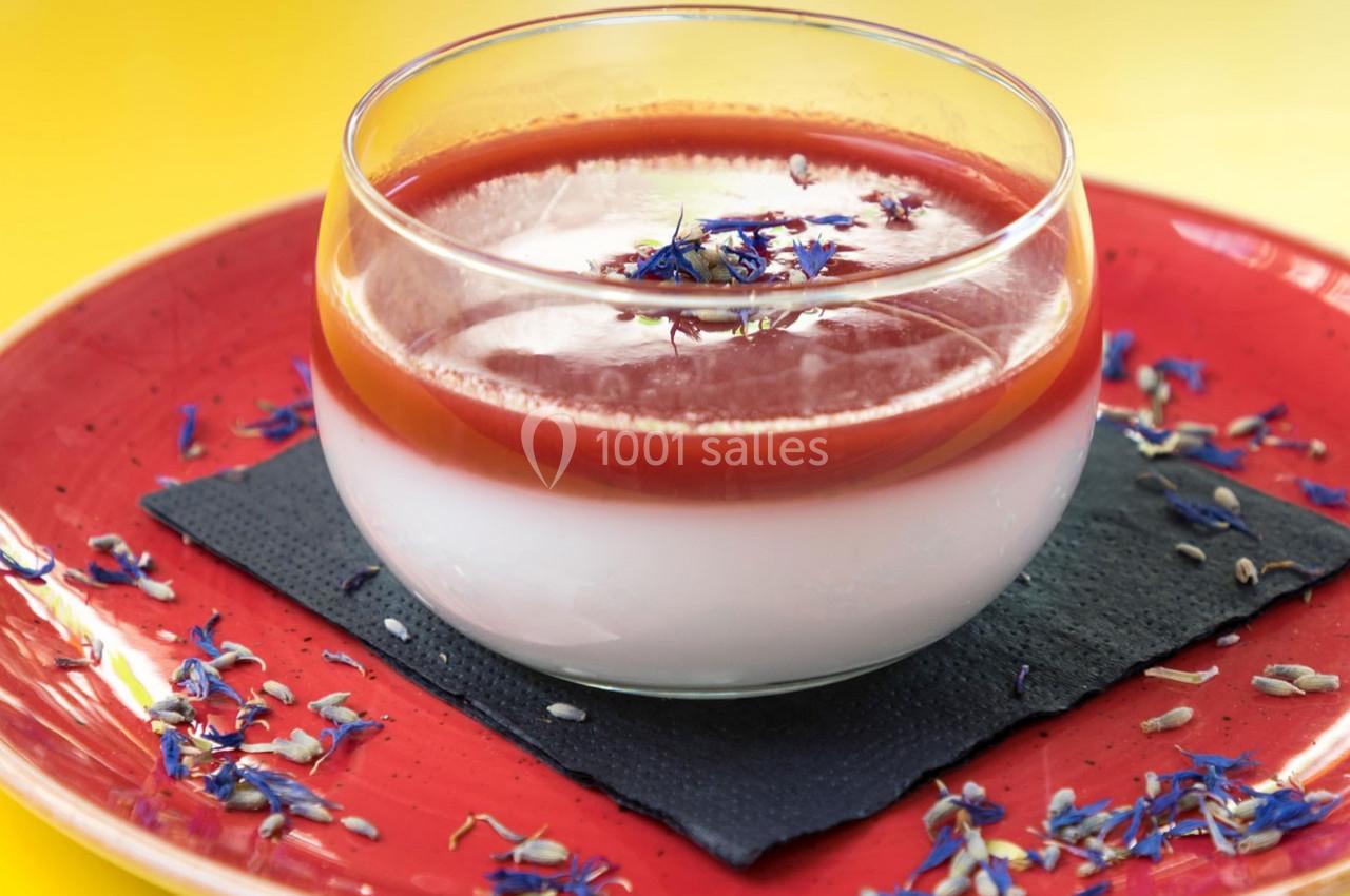 Dessert en verrine avec une couche blanche, un coulis rouge, décoré de fleurs comestibles, posé sur une assiette rouge.