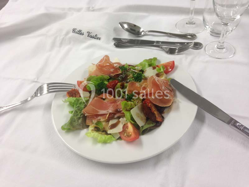 Assiette de salade garnie de jambon cru, tomates, parmesan et légumes, posée sur une nappe blanche avec couverts.