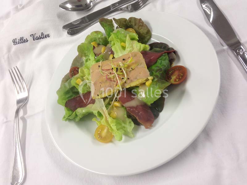 Assiette de salade composée avec foie gras, magret de canard fumé, tomates cerises et maïs, servie sur une nappe blanche.