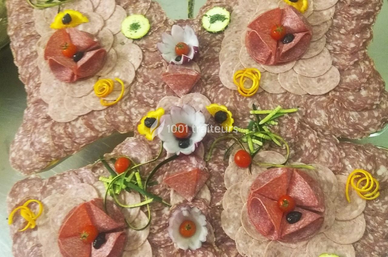 Plateau de charcuterie décoratif avec des tranches de saucisson, fleurs en légumes et garnitures colorées.