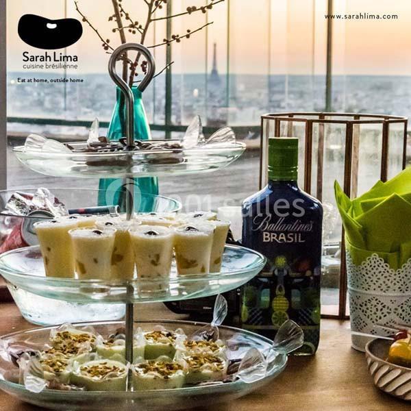 Plateau de desserts variés sur une table, avec une bouteille de liqueur et une vue panoramique sur une ville en arrière-plan.