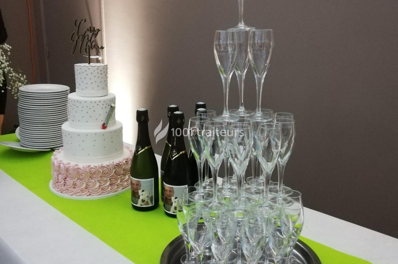 Table avec une pièce montée, des bouteilles de champagne, une pyramide de flûtes et une pile d'assiettes blanches.