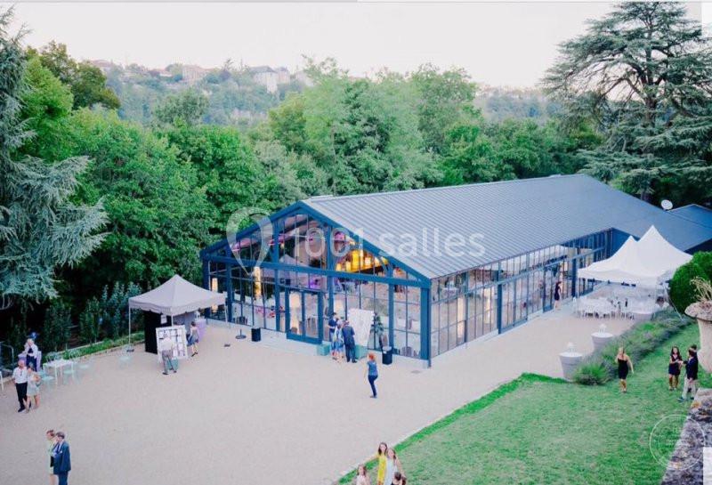 Vue d'une grande verrière moderne entourée de verdure, avec des invités et des tentes blanches à l'extérieur.