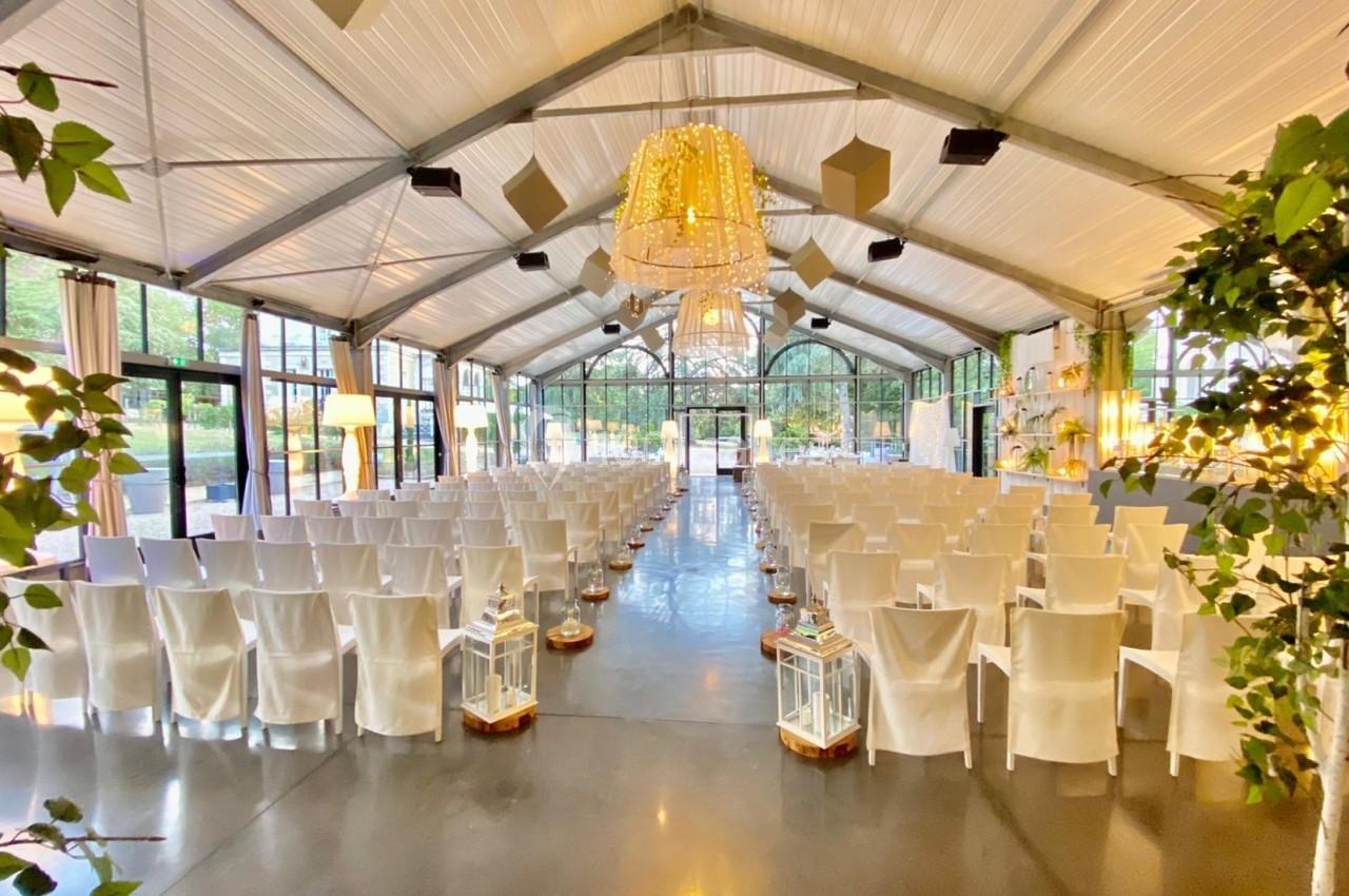 Salle lumineuse avec des rangées de chaises blanches, lanternes décoratives au sol et grand lustre central.