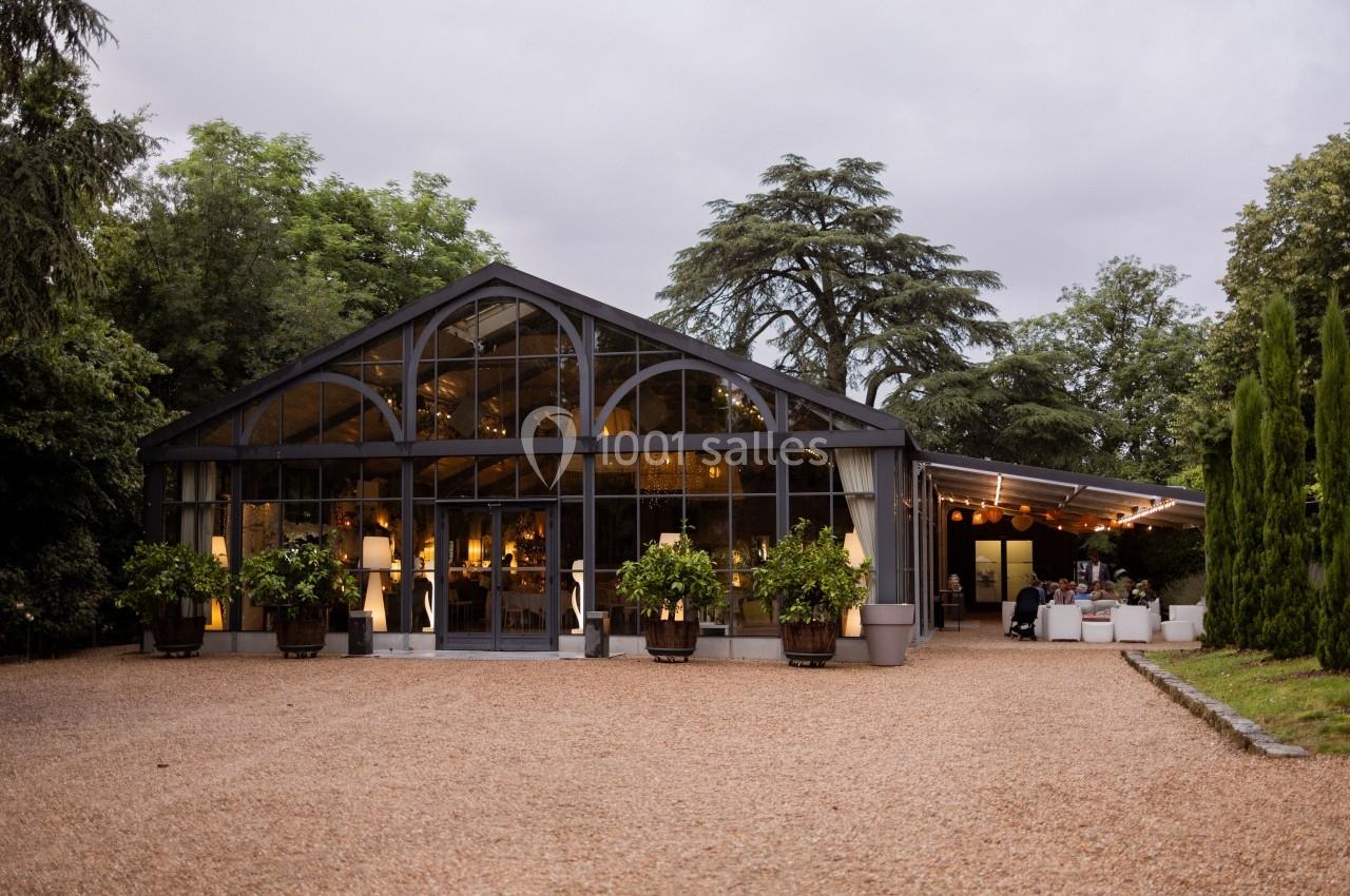 Serre moderne avec façade vitrée entourée de végétation, éclairée de l'intérieur, sur une allée de gravier.