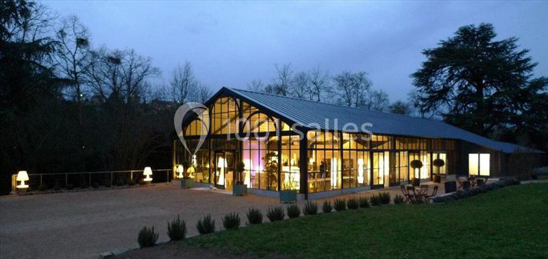 Serre moderne vitrée éclairée de nuit, entourée d'arbres et d'un espace extérieur aménagé avec des tables et des chaises.