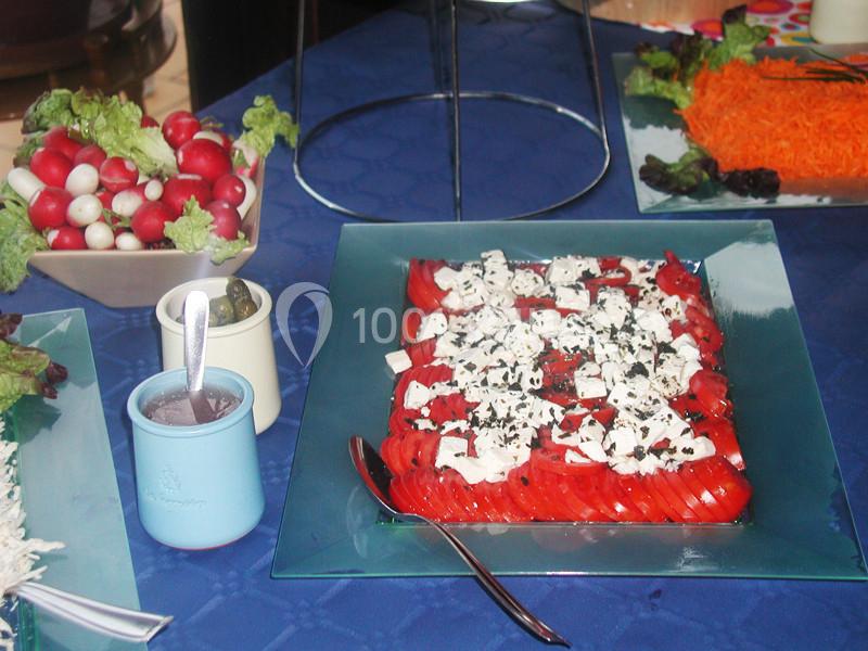 Assiette de tomates et fromage frais parsemée d'herbes, entourée de radis, carottes râpées et condiments sur une nappe bleue.