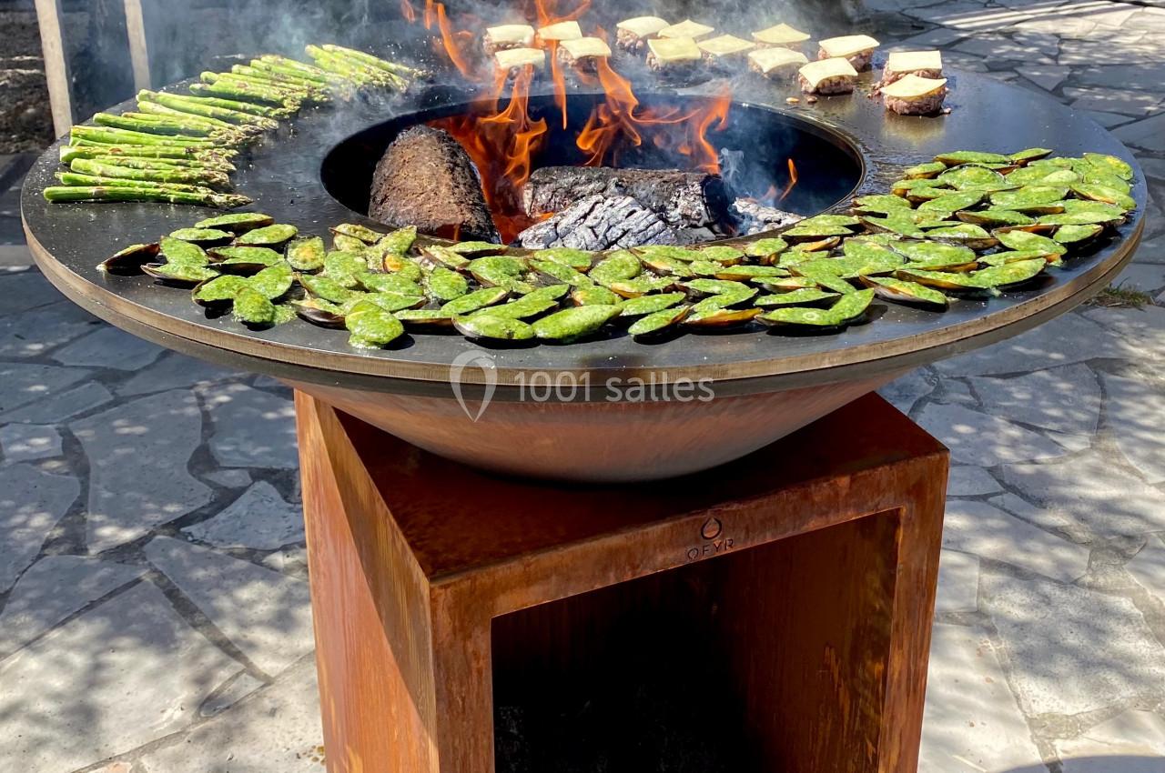 Grill circulaire avec des légumes verts et des aliments en cuisson sur un feu de bois dans un espace extérieur ensoleillé.