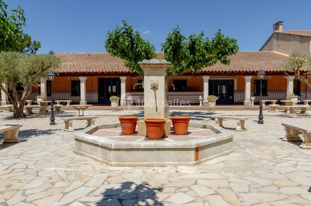 Place pavée avec une fontaine centrale entourée de pots en terre cuite, bancs en pierre et bâtiments à arcades.
