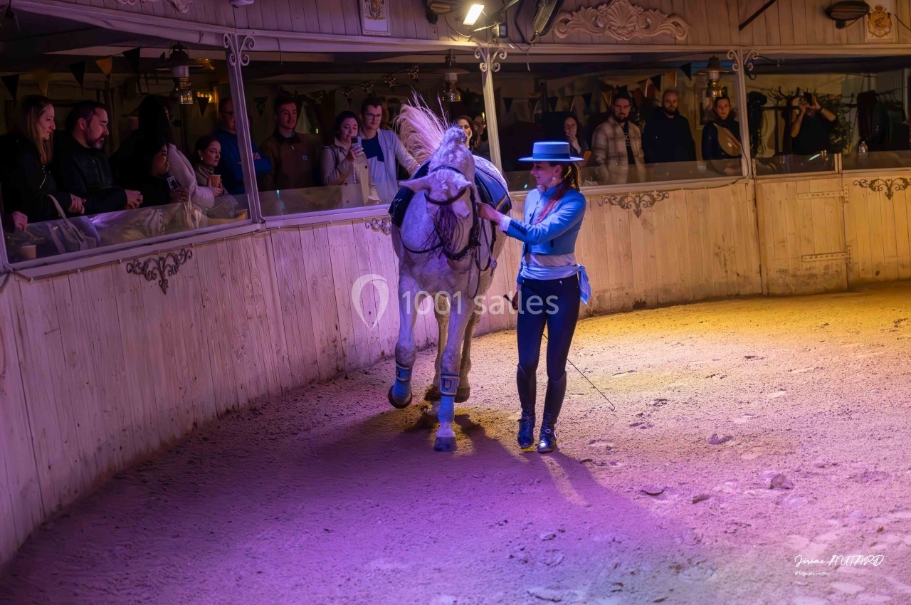 Une cavalière en tenue traditionnelle guide un cheval blanc dans une arène devant un public attentif.