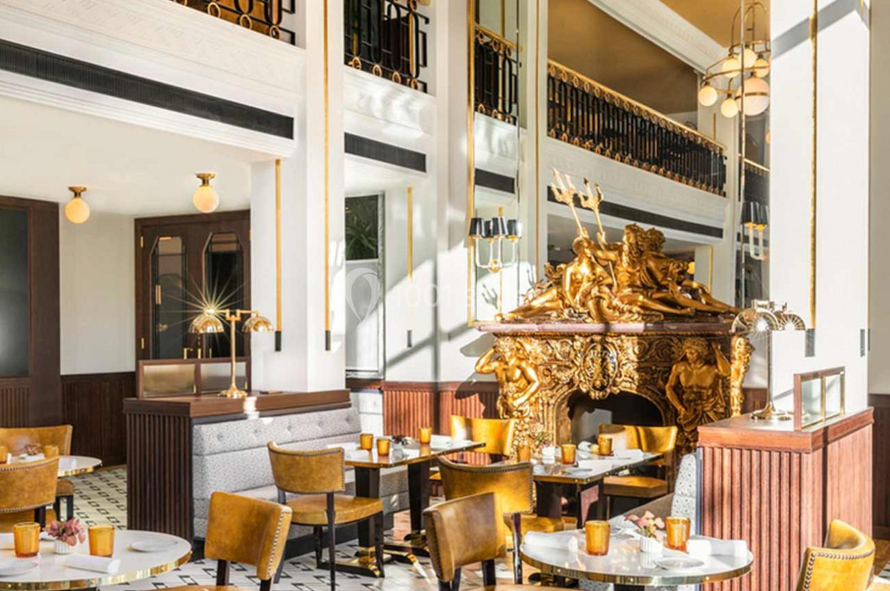 Salle de restaurant élégante avec mobilier doré, cheminée ornée de sculptures et lumière naturelle abondante.