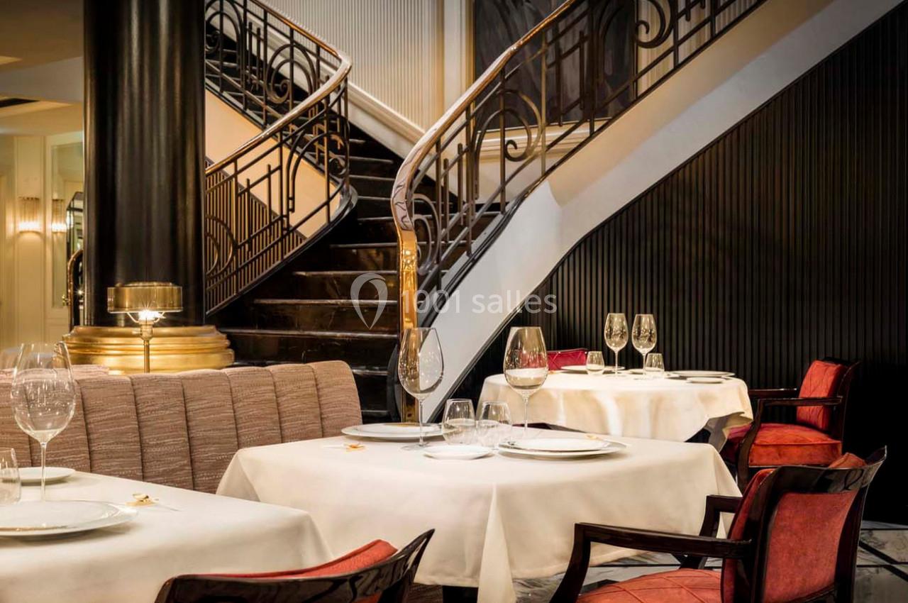 Salle de restaurant élégante avec tables dressées, chaises en bois et escalier en colimaçon en arrière-plan.