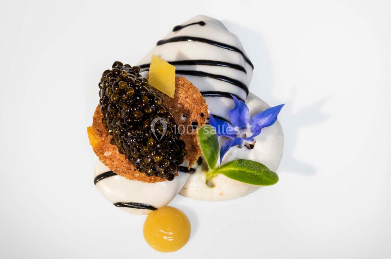 Dessert raffiné avec mousse blanche, caviar, biscuit, fleur comestible bleue et sauce jaune sur assiette blanche.