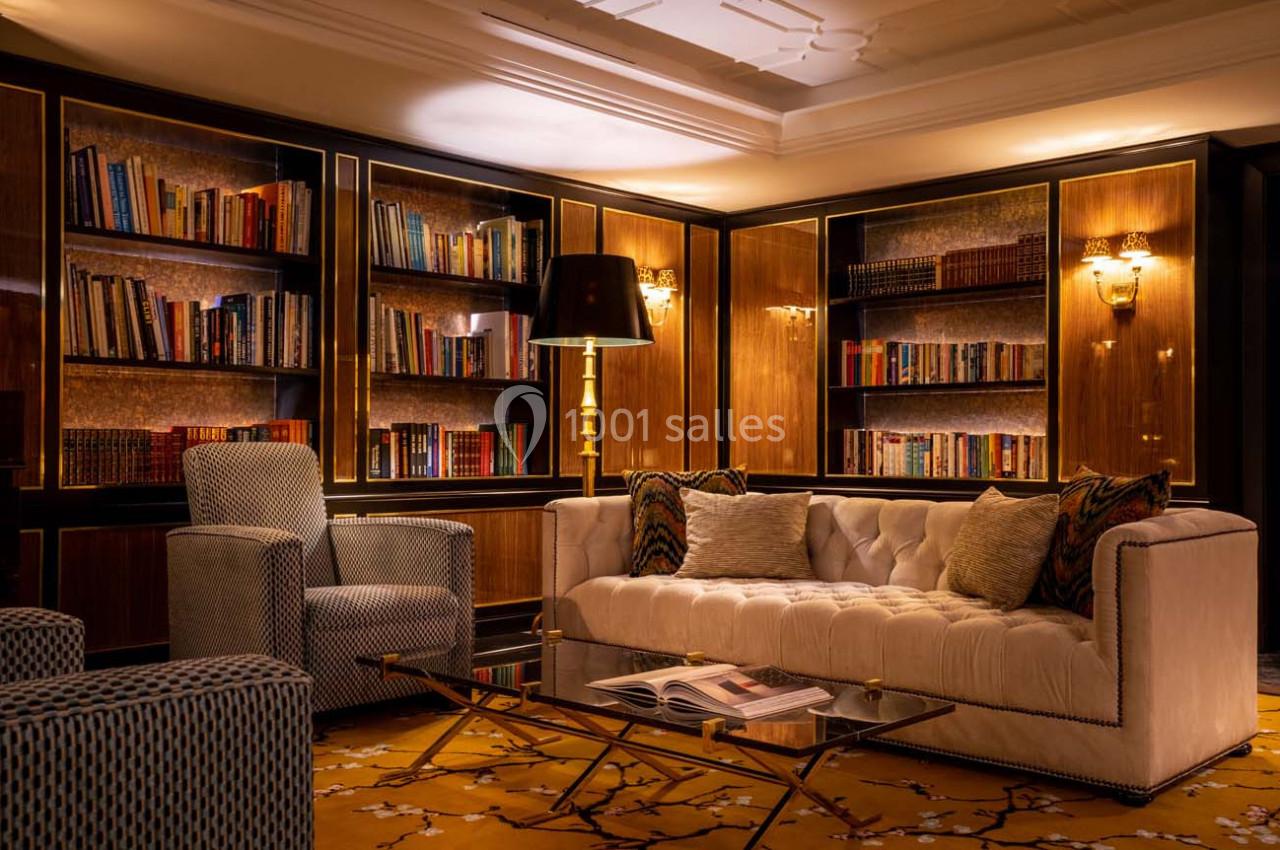 Salon élégant avec canapé capitonné, fauteuils, table basse en verre, lampadaire et bibliothèque en bois éclairée.