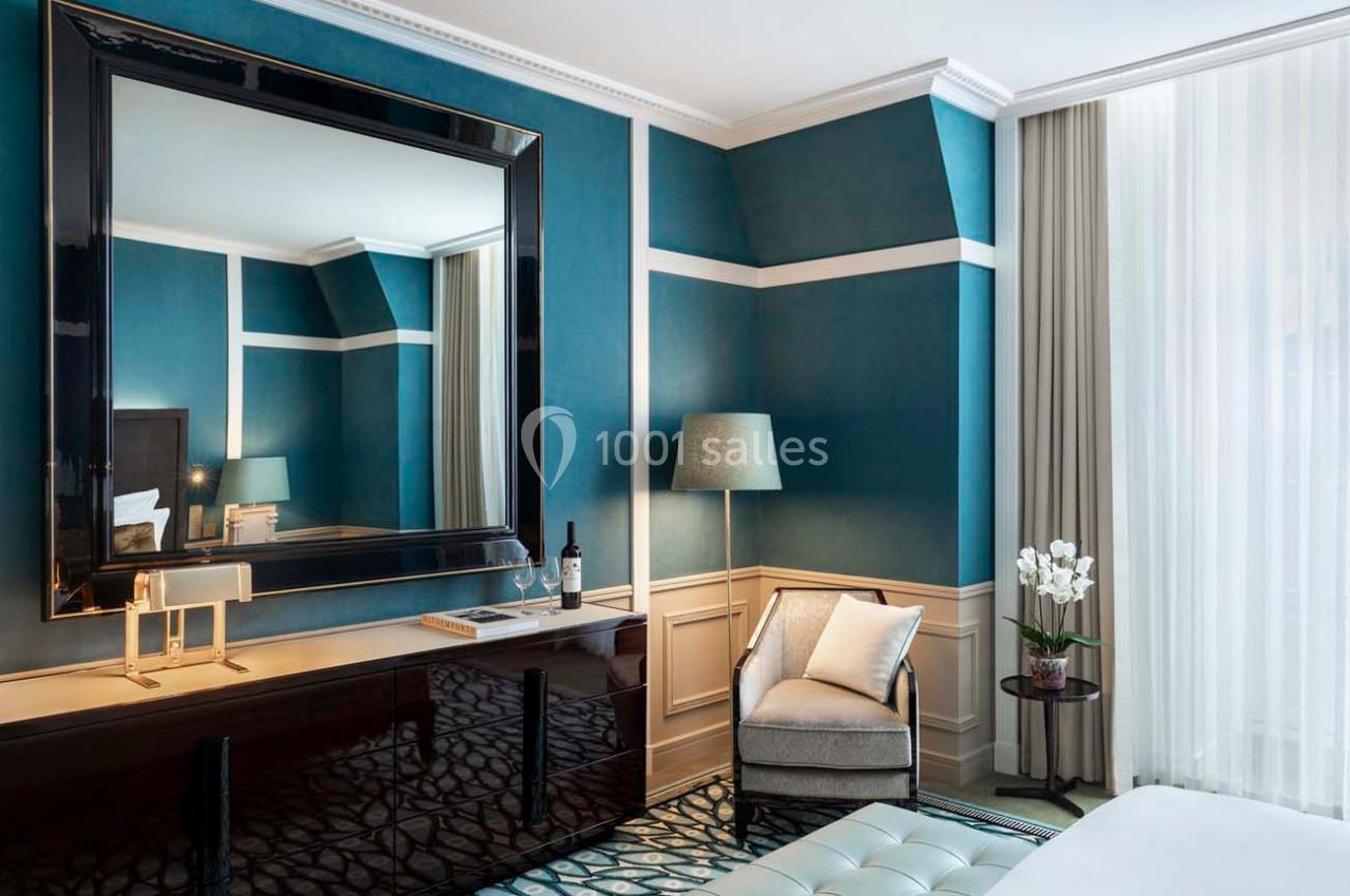 Chambre élégante avec murs bleus, grand miroir, fauteuil, lampe, rideaux translucides et décoration florale.