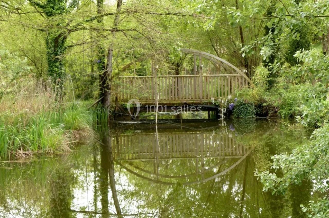 Passerelle en bois traversant un étang entouré de végétation dense et reflétée dans l'eau calme.