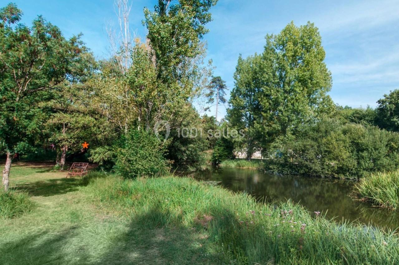 Paysage naturel avec un étang entouré d'arbres, d'herbes hautes et d'un banc en bois à l'ombre.