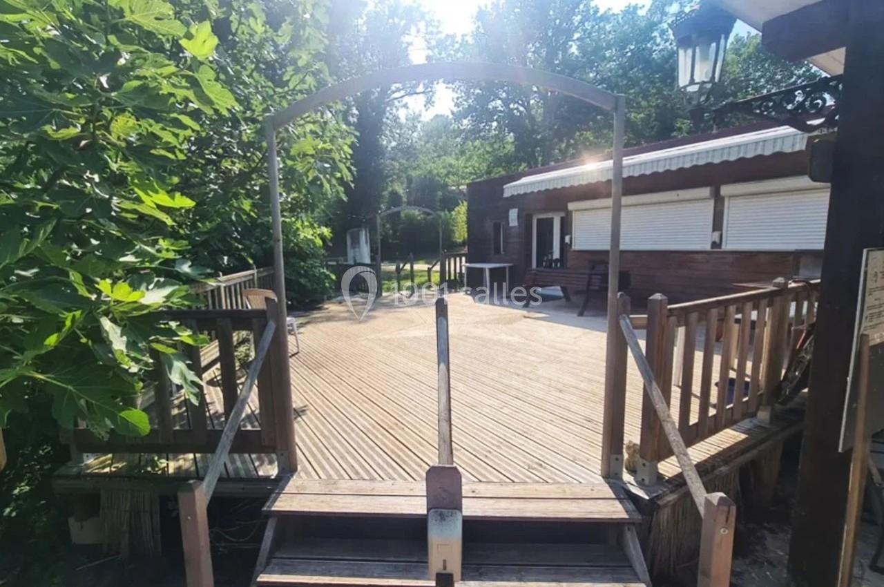 Passerelle en bois menant à une terrasse avec une maison en arrière-plan, entourée de végétation.