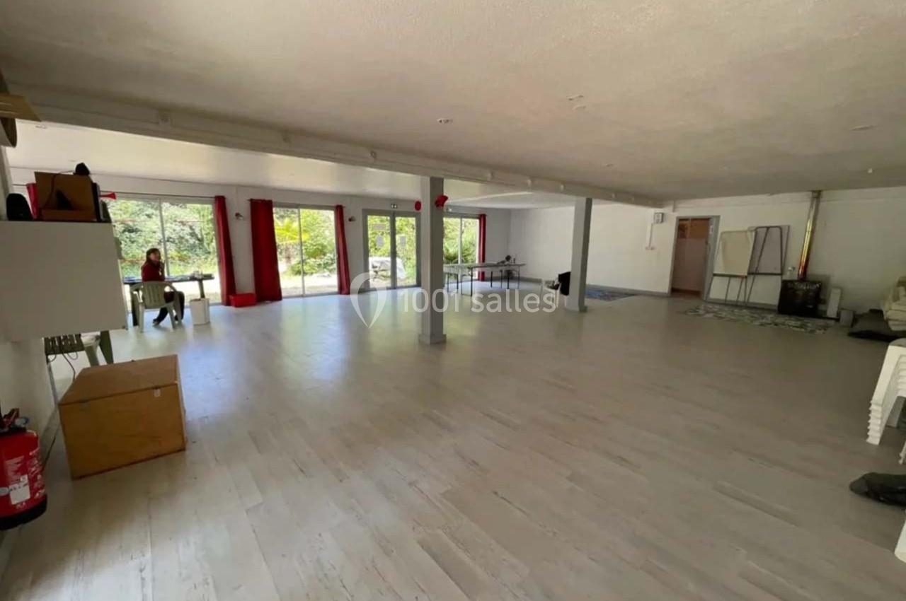 Salle spacieuse avec parquet clair, grandes baies vitrées donnant sur un jardin, et quelques meubles dispersés.