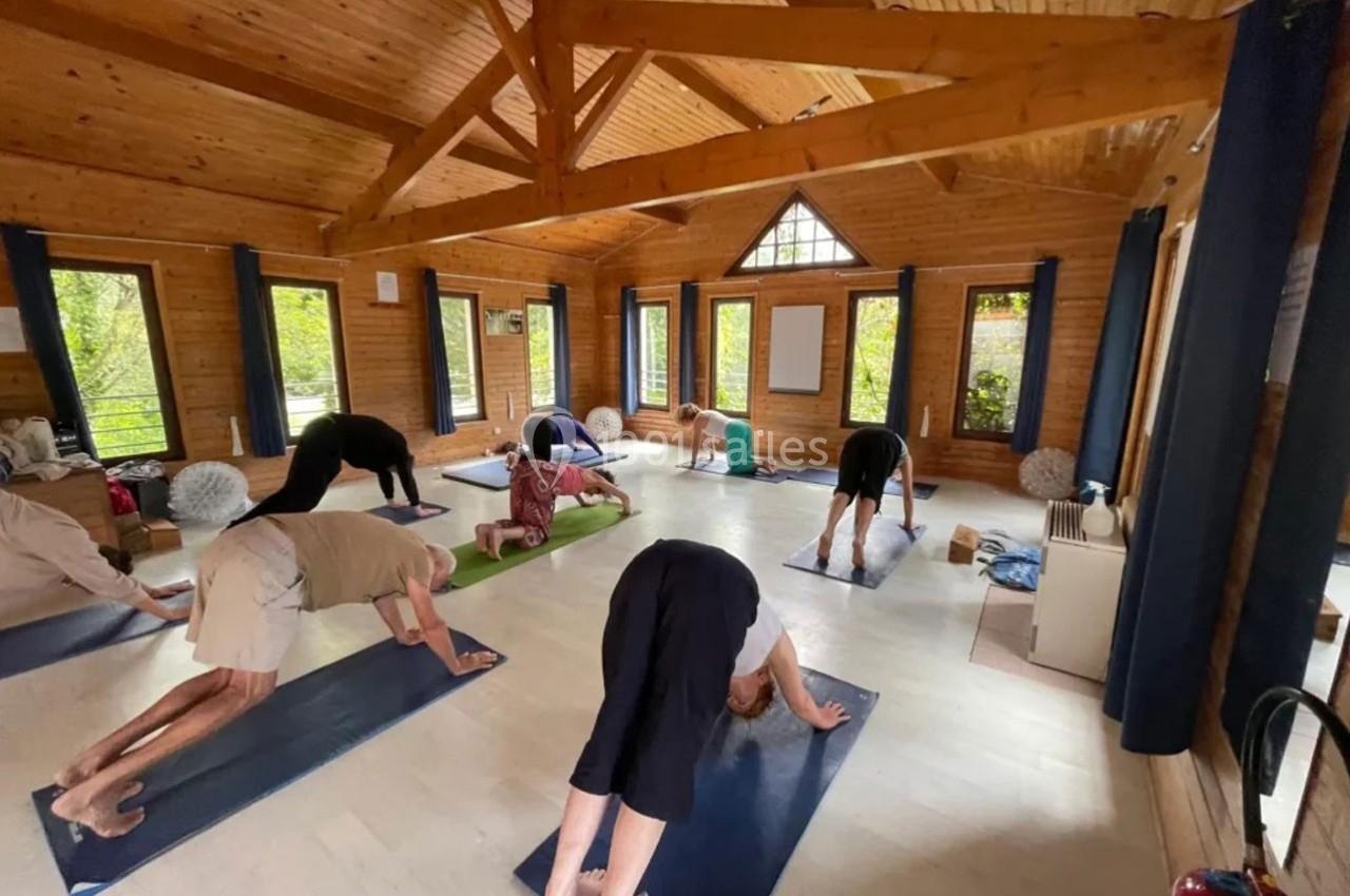 Personnes pratiquant le yoga dans une salle en bois lumineuse, avec des tapis disposés au sol.