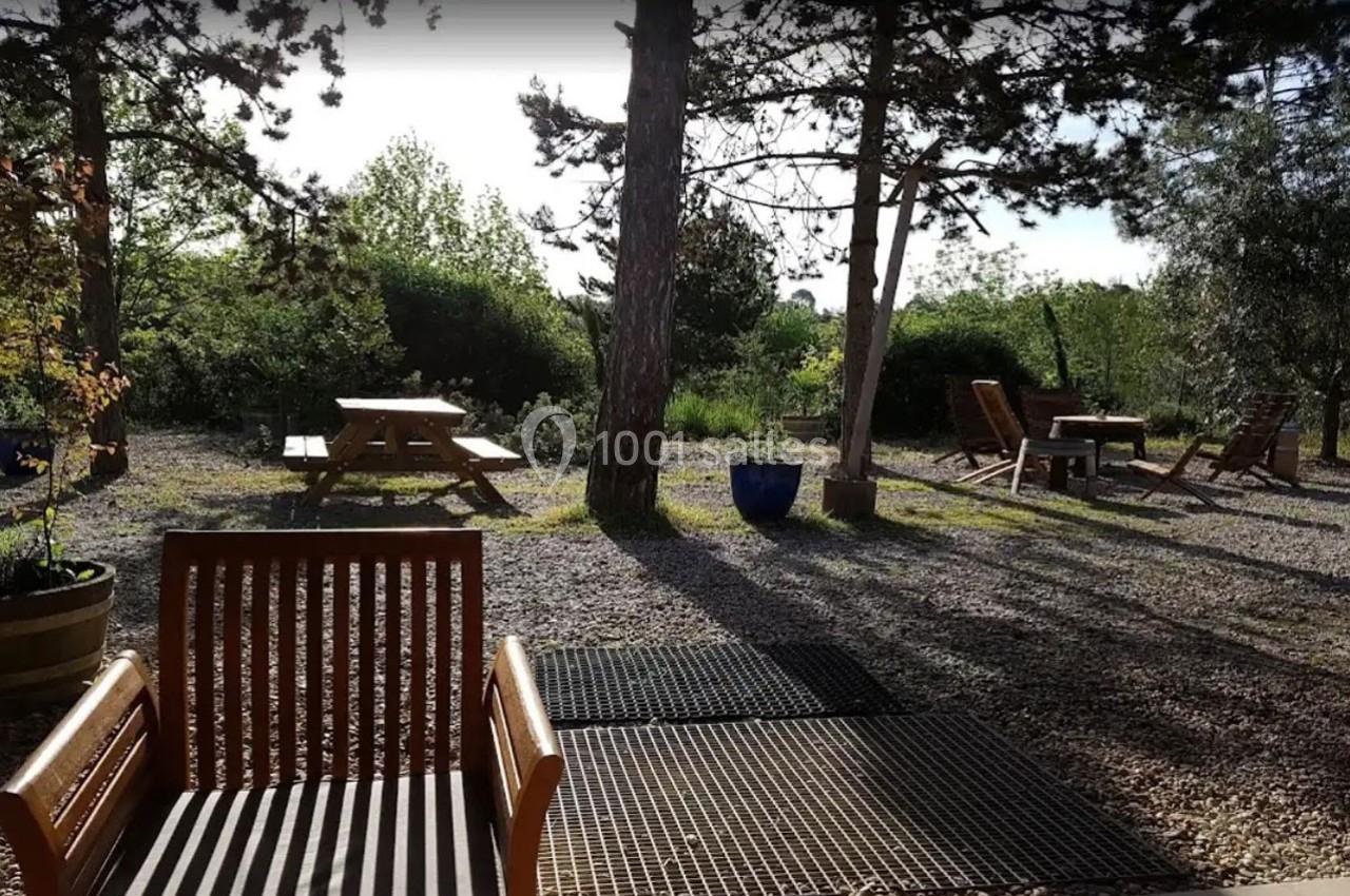 Espace extérieur avec des bancs et des tables en bois, entouré d'arbres et de végétation sous une lumière naturelle.