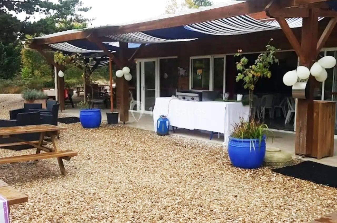 Terrasse avec tables en bois, pots bleus et décorations, devant un bâtiment avec grandes baies vitrées et auvent rayé.