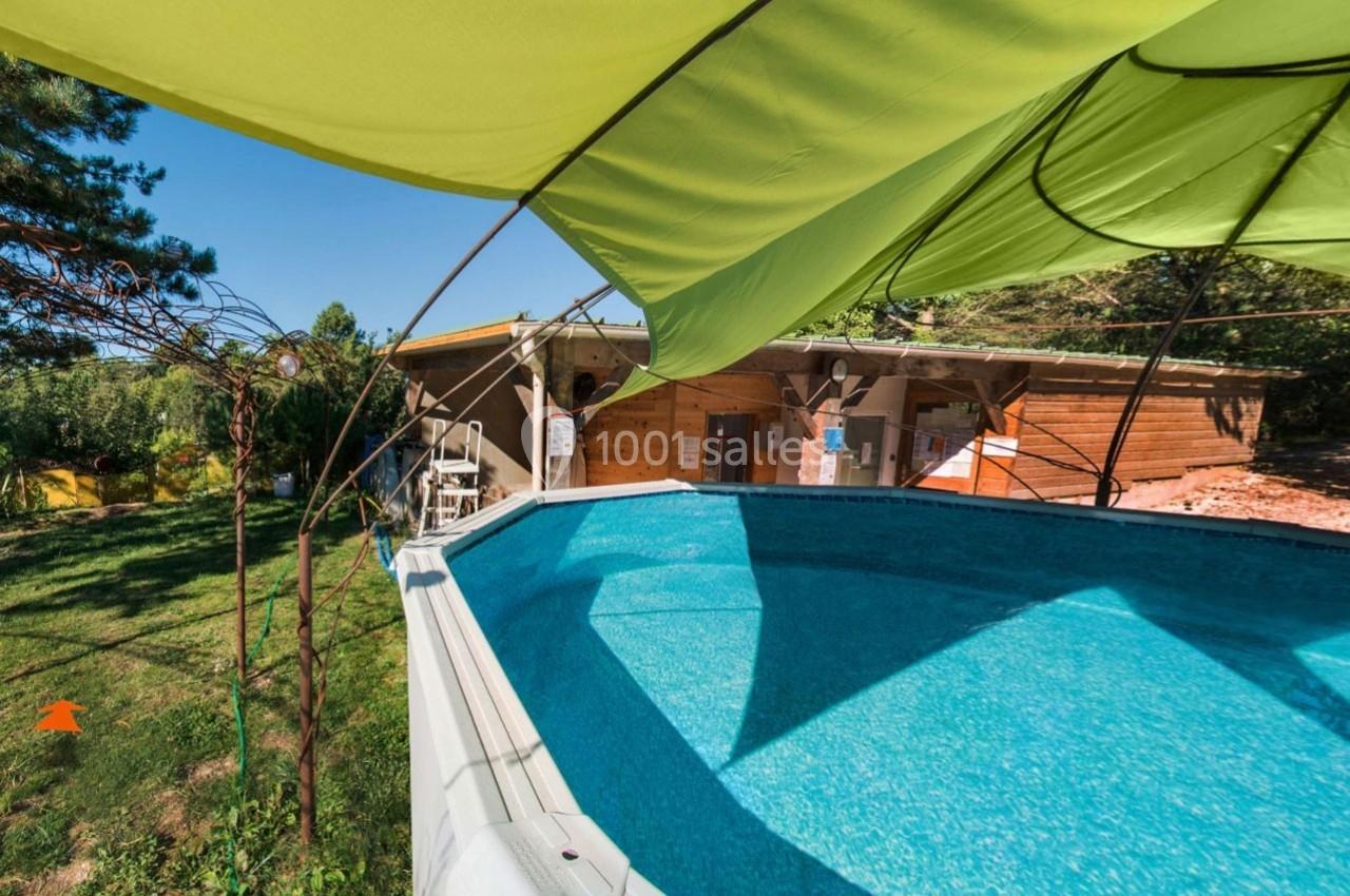 Piscine hors-sol entourée de verdure, avec un abri en bois et un grand parasol vert en arrière-plan.