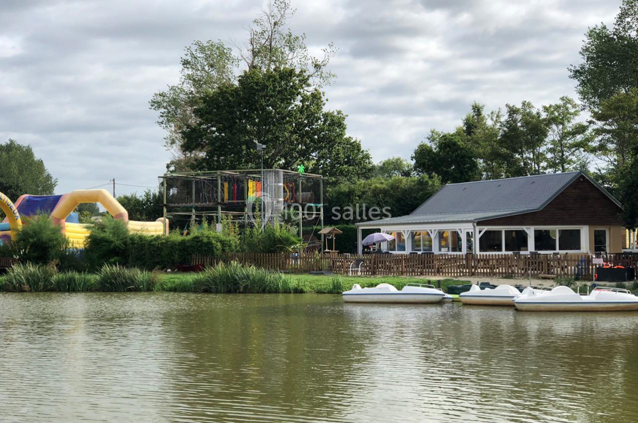 Plan d'eau avec pédalos au premier plan, structure de jeux et bâtiment en bois entourés de végétation en arrière-plan.