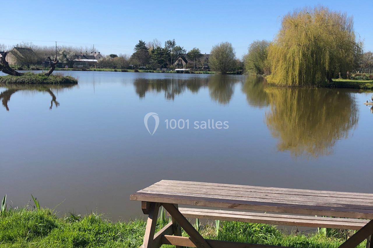 Vue d'un étang calme entouré d'arbres sous un ciel bleu, avec une table de pique-nique en bois au premier plan.
