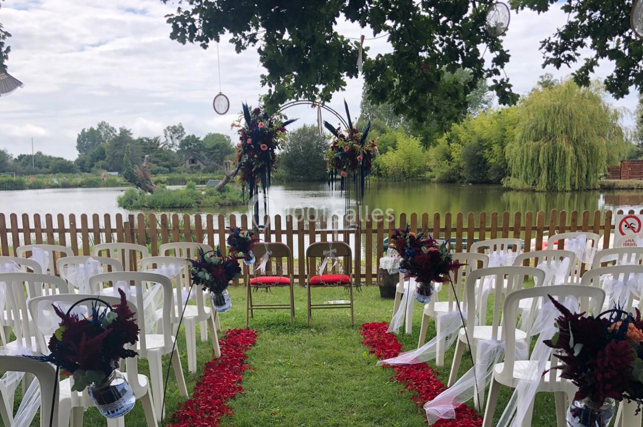 Chaises blanches alignées devant une arche fleurie, disposées pour une cérémonie en plein air près d'un étang.