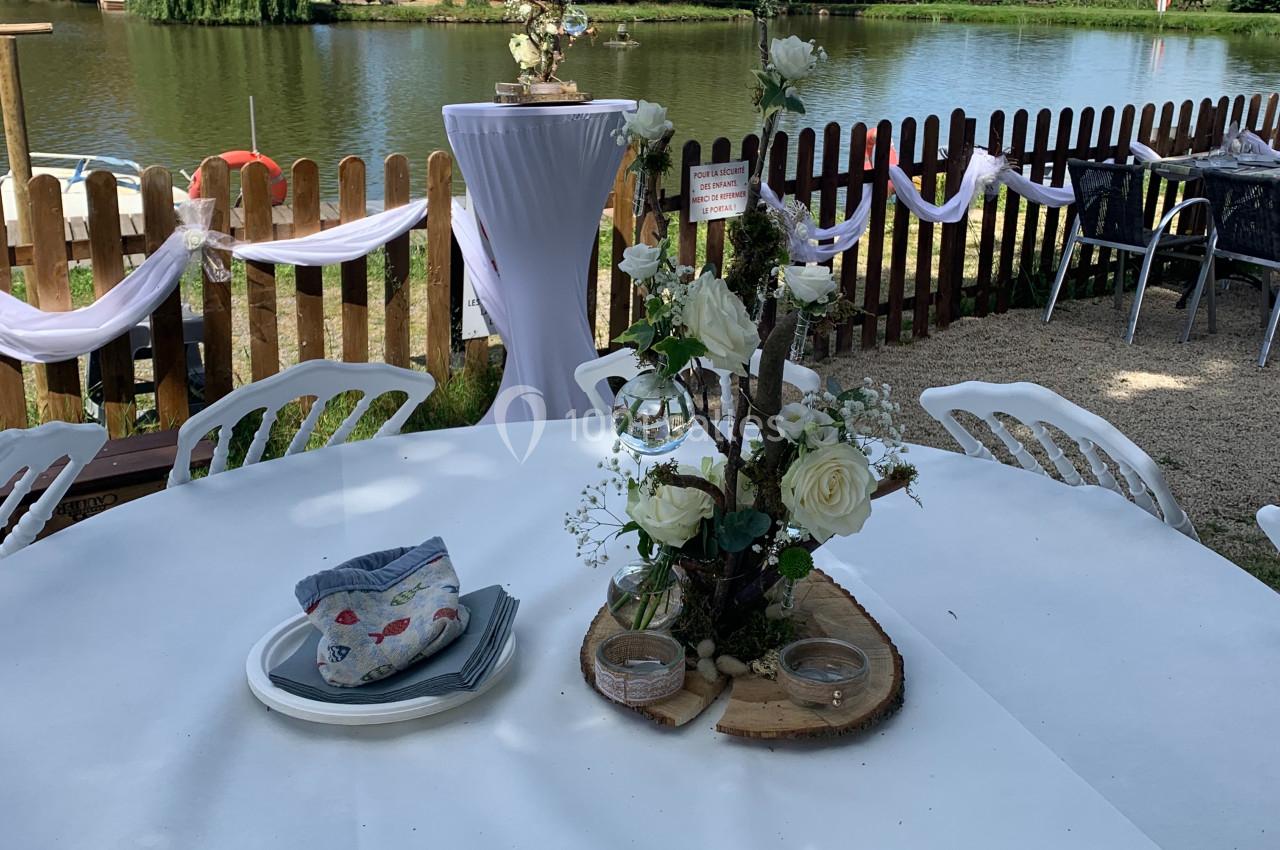 Table décorée avec des fleurs blanches et des rondins de bois, près d'un lac entouré de verdure et d'une clôture en bois.