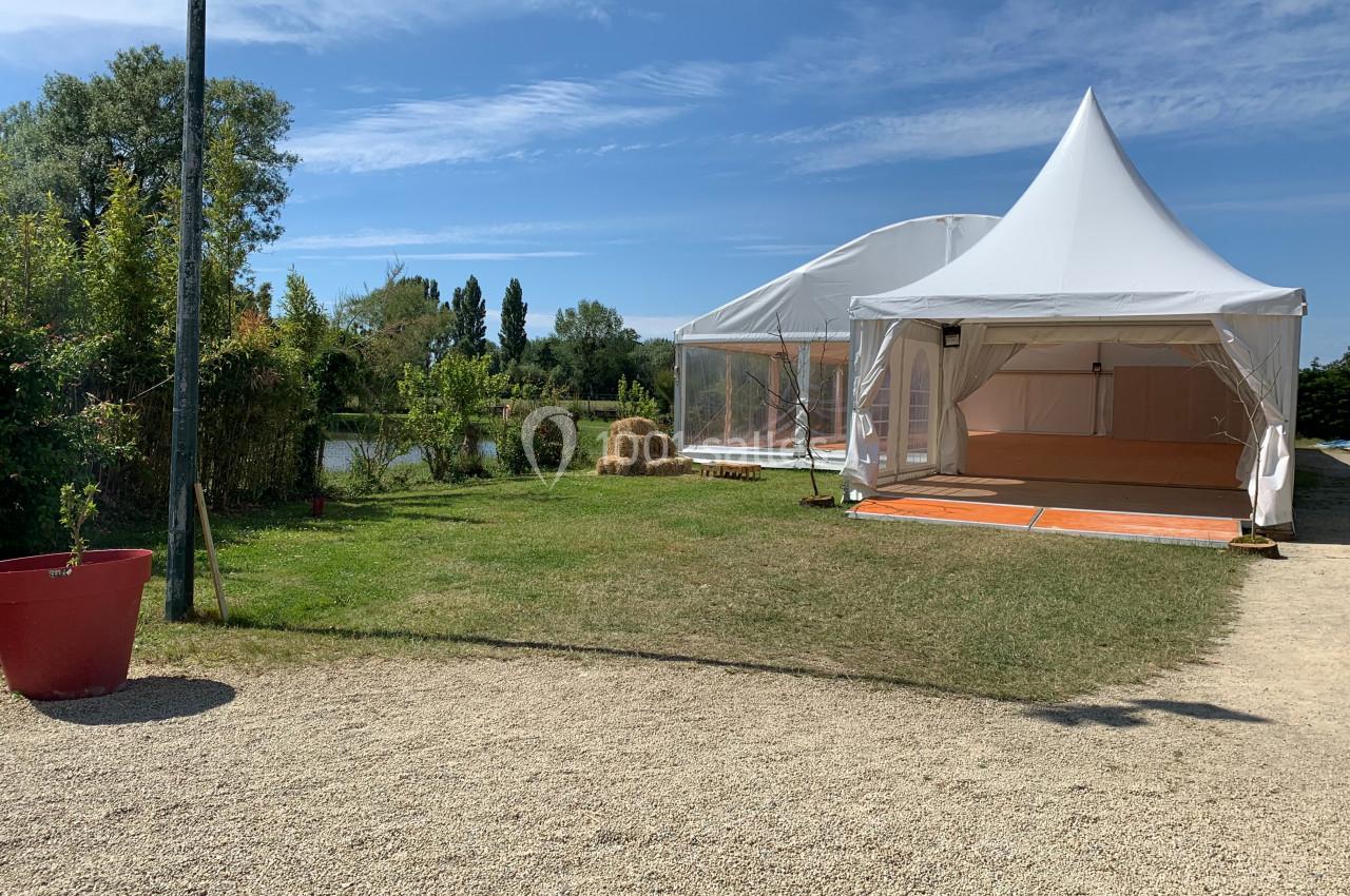 Deux grandes tentes blanches installées sur une pelouse près d'un étang, entourées de végétation et de bottes de paille.